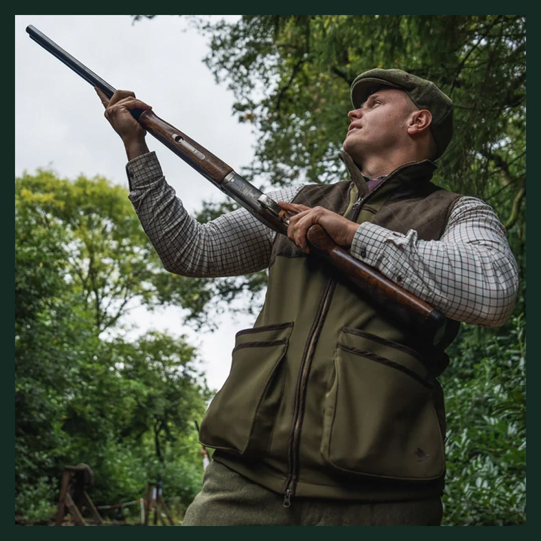 Man wearing a traditional shooting vest, checked shirt and flat cap while clay shooting in a woodland setting.