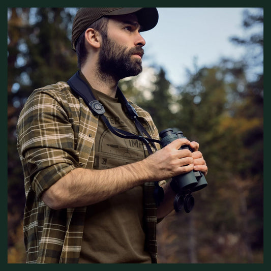 Man wearing a green checked country shirt and cap, holding binoculars while observing the surrounding woodland.