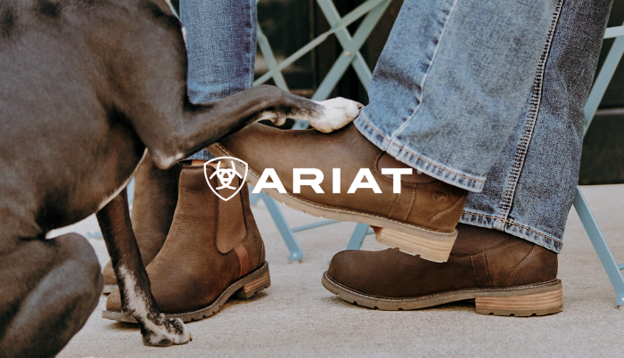 A person wearing Ariat Wexford boots with blue jeans, with a dog sitting beside them and placing its paw on the boot. The Ariat logo is overlaid in the centre.