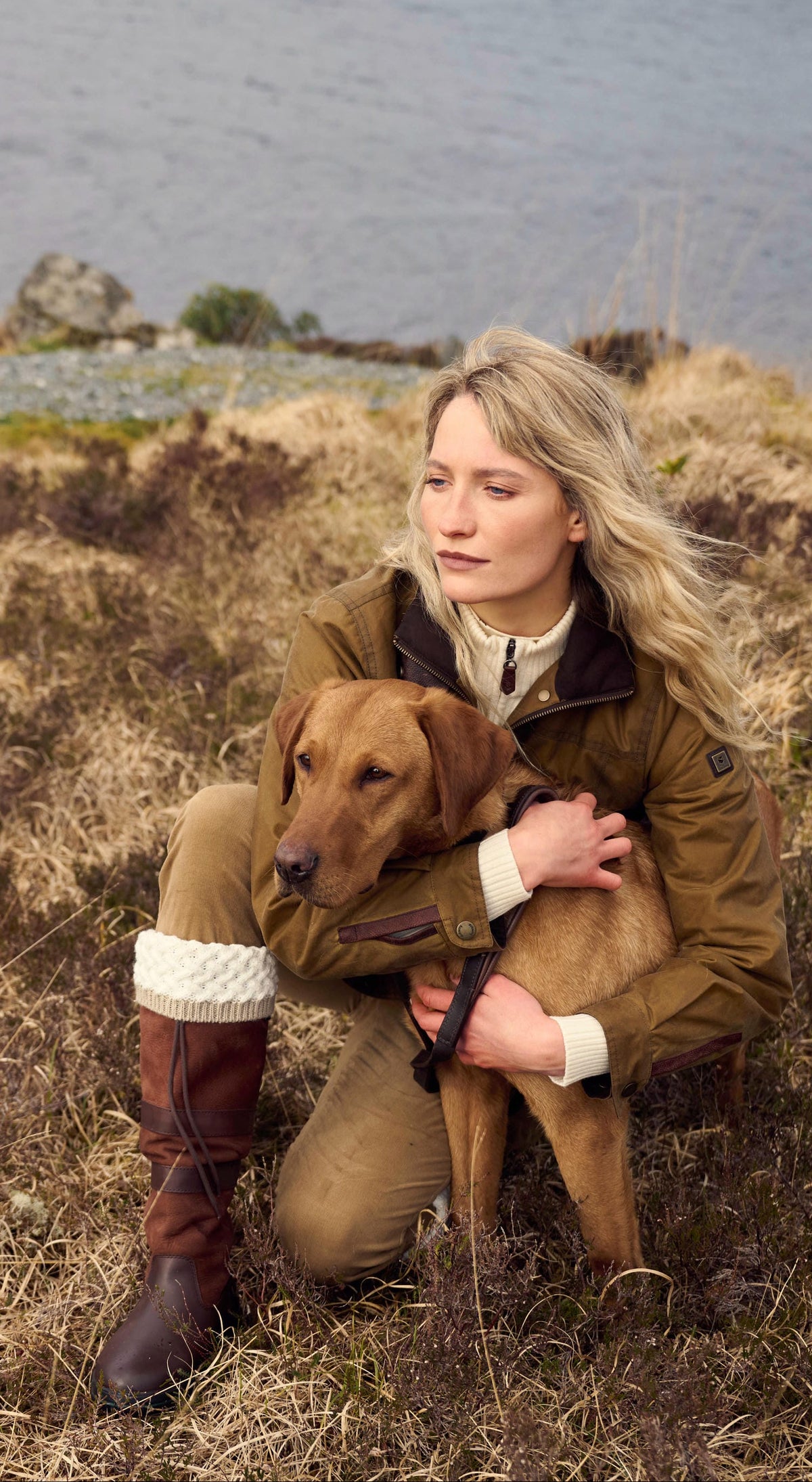 A woman crouching in a grassy field by the water, holding a golden retriever dog. She is wearing a Dubarry Mountrath jacket, beige trousers, and Dubarry Galway boots.