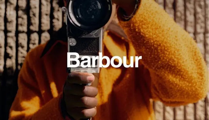 A person in a textured mustard yellow sweater holds a vintage film camera up to their face, with the word 'Barbour' overlaid in large white text across the center.