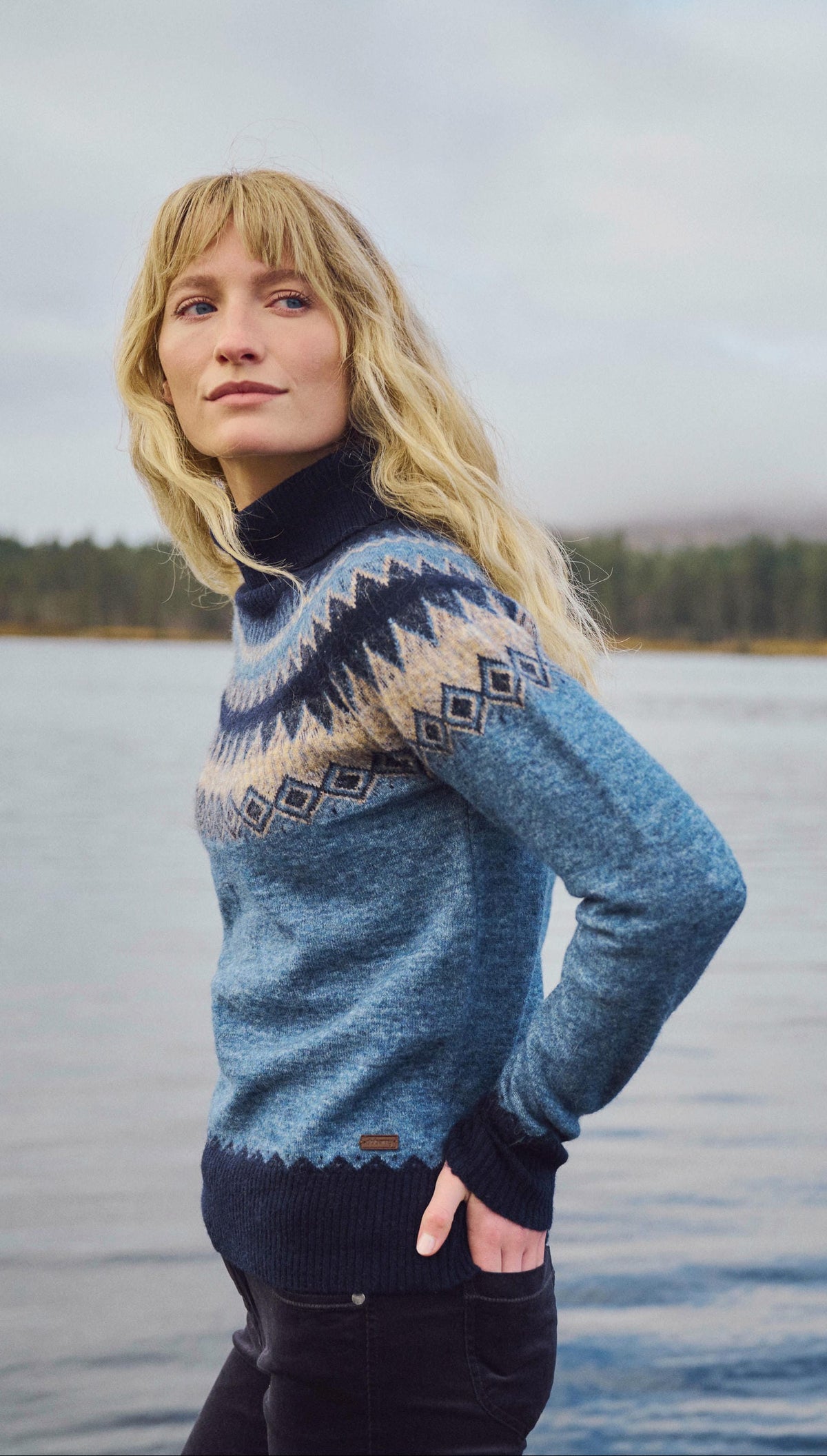 Woman wearing a Navy Dubarry Devlin knit jumper beside a loch, with mountains and woodland in the background.