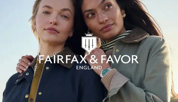 Two women outdoors with the Fairfax & Favor England logo overlaid in the center. One wears a navy jacket with gold-tone hardware, and the other wears an olive green jacket over a striped top.