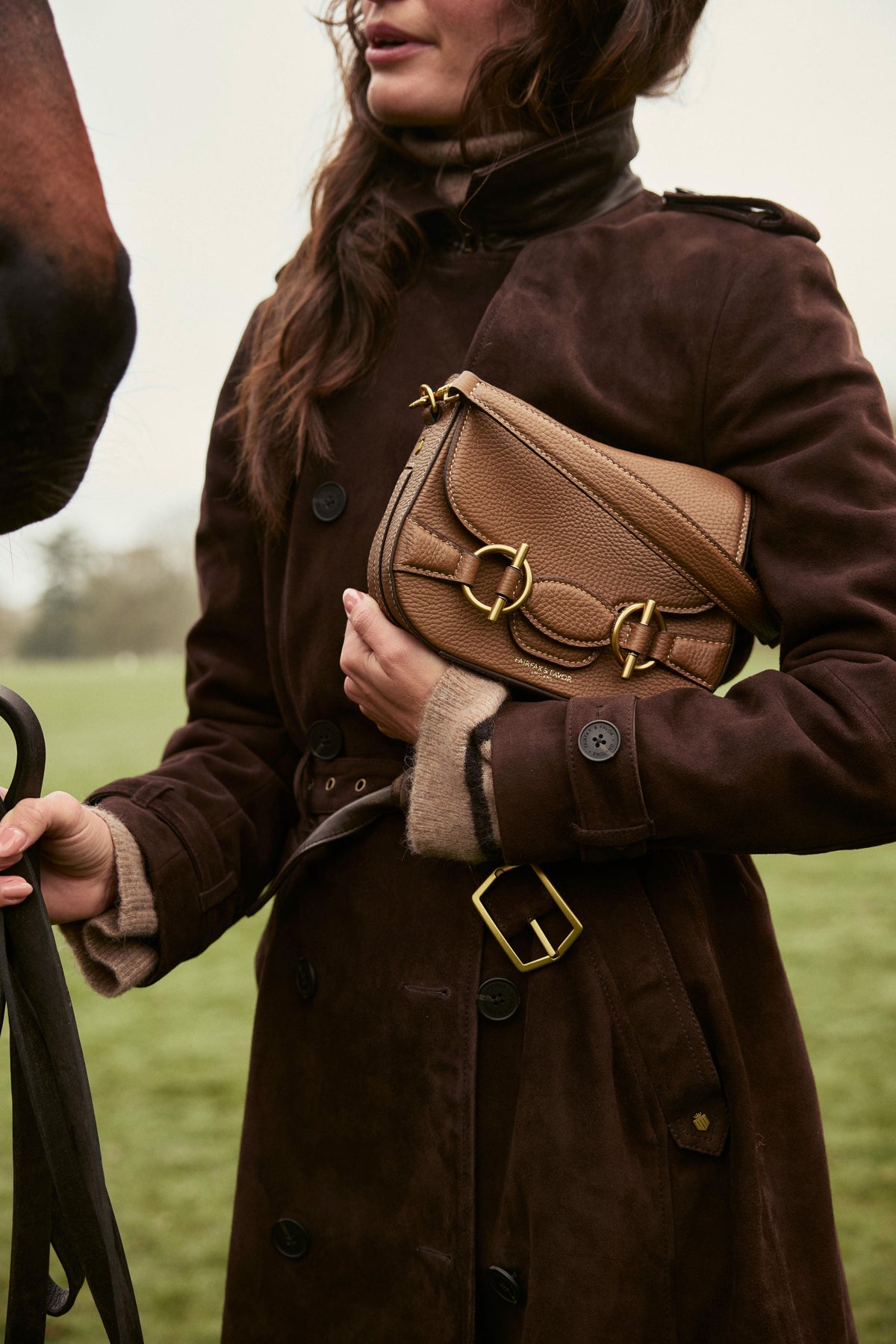 Woman carrying a tan Fairfax & Favor Connie Bag, styled with a brown belted country coat beside a horse.