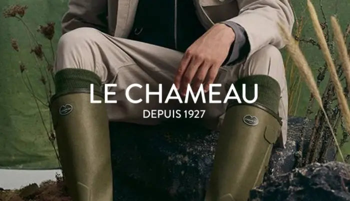 A person seated outdoors wearing olive green Le Chameau wellington boots, with a beige jacket and trousers visible. Large white text across the center reads 'LE CHAMEAU DEPUIS 1927'.
