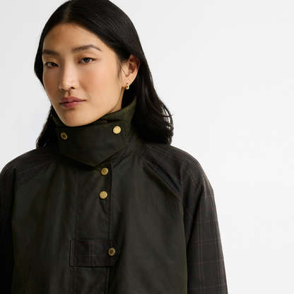 A close-up of a model wearing the Barbour Kirklees Checked Waxed Jacket. Featuring a fern green body with check detailing on the sleeves and brass snap fastenings. 