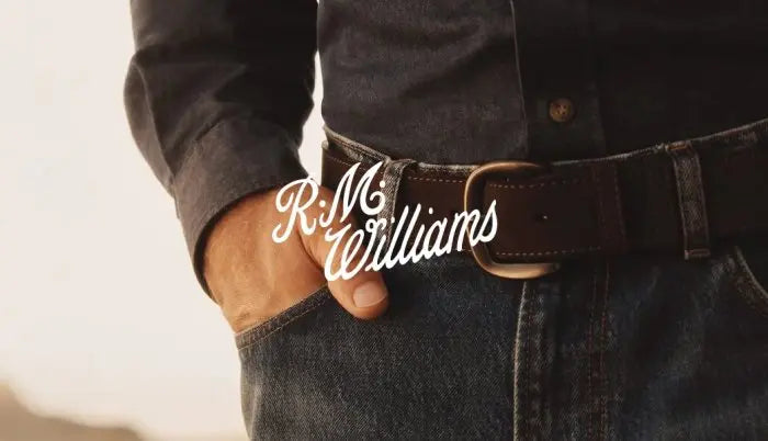 A close-up lifestyle shot of a person wearing a dark brown leather belt with a brass buckle, paired with a dark button-down shirt and blue jeans. The brand name 'R.M. Williams'.
