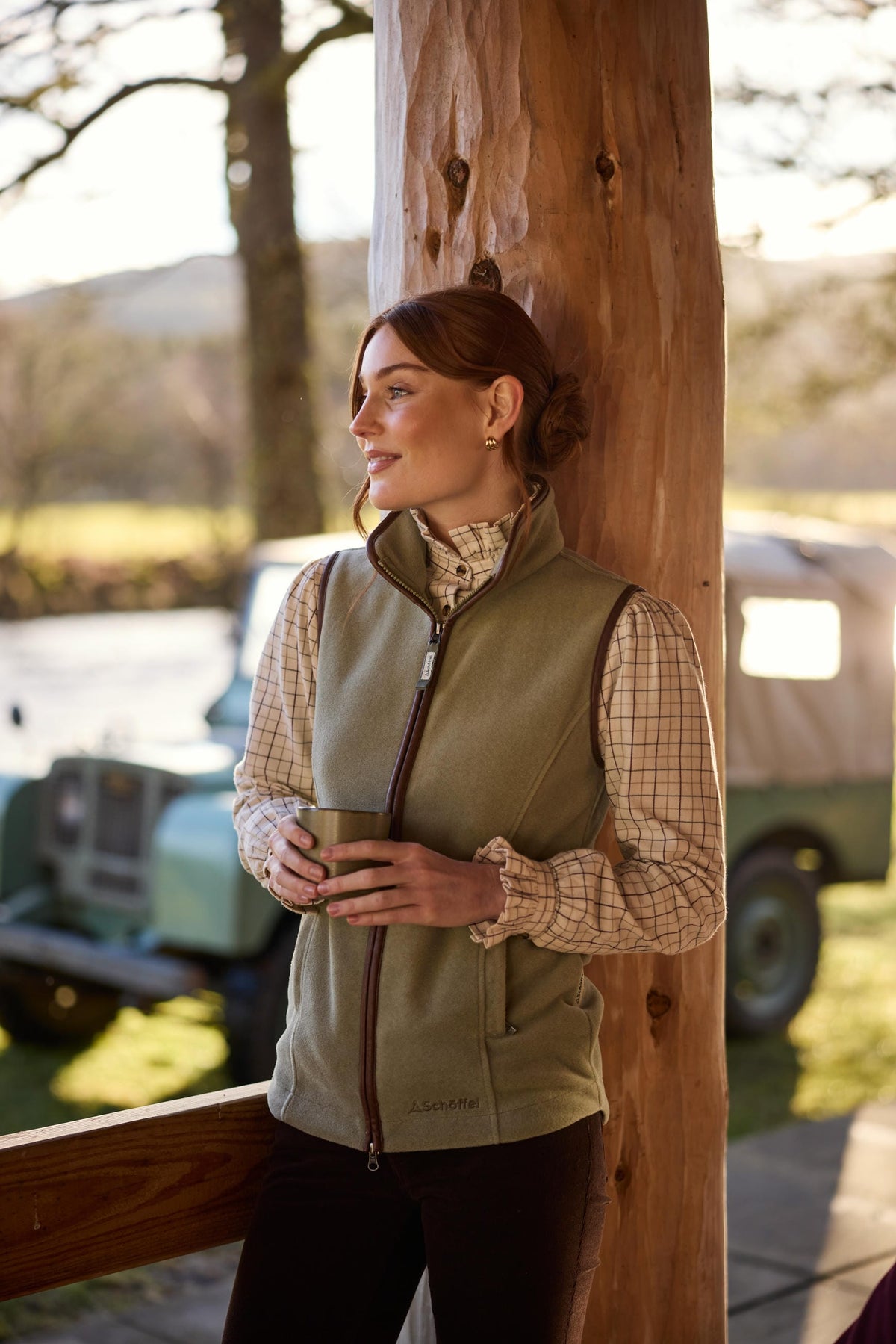 Woman wearing a Schöffel Lyndon fleece gilet in Sage Green layered over a checked country shirt, holding a mug beside a classic Land Rover in the countryside.
